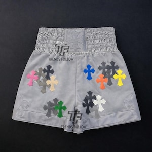 Puede incluir: Pantalones cortos deportivos grises con un patrón de cruces. Los pantalones cortos tienen una cintura elástica gris y presentan múltiples cruces de colores, incluyendo rosa, azul, naranja y amarillo. El texto "TRENDS FOLLOW" es visible.