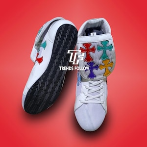 以下が含まれることがあります： カラフルな十字架のデザインが施された白いスニーカー。靴は白いメッシュのアッパー、黒いソール、十字架の装飾が付いたストラップが付いています。「TRENDS FOLLOW」の文字が見えます。