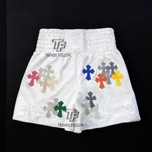 Puede incluir: Pantalones cortos deportivos blancos con múltiples aplicaciones en forma de cruz de varios colores. Los pantalones cortos tienen una cintura elástica blanca y el texto "TRENDS FOLLOW" es visible. Los pantalones cortos están hechos de un material brillante.