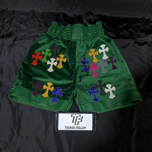 Puede incluir: Pantalones cortos de boxeo de satén verde esmeralda con aplicaciones de cruces multicolores. Los pantalones cortos tienen una cintura elástica ancha y están bordados con cruces de varios colores. El texto "TRENDS FOLLOW" es visible en un teléfono.