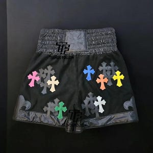 Op de afbeelding: Zwarte sportbroek met meerdere kruisvormige patches in verschillende kleuren. De broek heeft een brede, elastische tailleband en een leren afwerking. De tekst "TRENDS FOLLOW" is op de broek gedrukt.