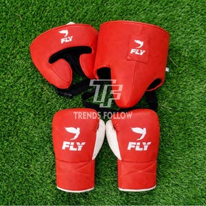 Puede incluir: Juego de guantes y casco de boxeo rojos con detalles blancos. Los guantes y el casco tienen la palabra "FLY" y un logotipo de pájaro. El conjunto se muestra sobre una superficie de hierba verde.