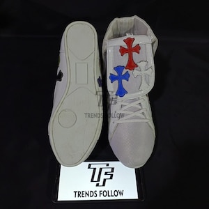 Puede incluir: Zapatillas blancas con cruces decorativas rojas, azules y blancas. Las zapatillas tienen un diseño texturizado y cierre con cordones. Se ve la suela de un zapato y el otro tiene una solapa con el diseño de la cruz.