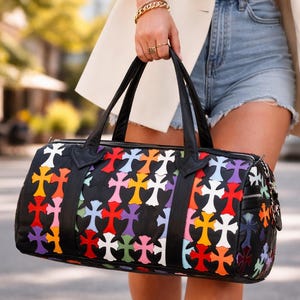 May include: Black duffel bag with a colorful cross pattern. The bag features a black leather trim, handles, and zipper. The crosses are in various colors, including red, yellow, purple, and white. The bag is held by a person wearing a white blazer and denim shorts.
