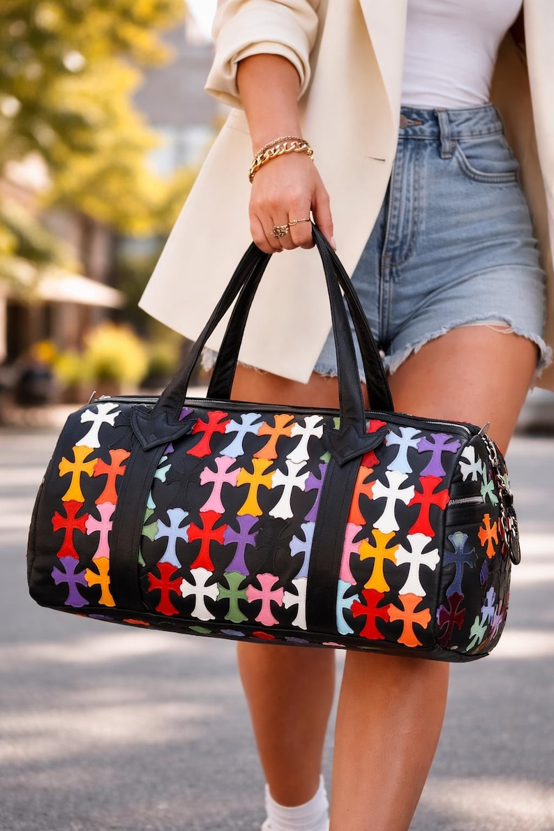 May include: Black duffel bag with a colorful cross pattern. The bag features a black leather trim, handles, and zipper. The crosses are in various colors, including red, yellow, purple, and white. The bag is held by a person wearing a white blazer and denim shorts.