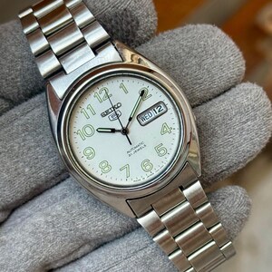 May include: A silver-toned Seiko 5 automatic wristwatch with a white face. The watch features a day and date display, luminous hands and numerals, and a stainless steel band. The watch is displayed on a gray surface.