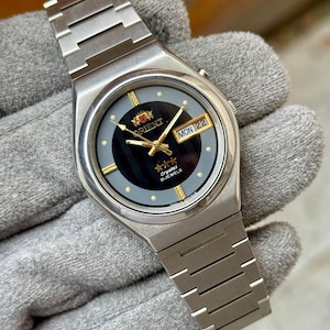 May include: A silver-toned stainless steel wristwatch featuring a round case and a matching link bracelet. The black dial displays gold-colored hands, hour markers, a day-date window at the three o'clock position, and the text Orient Crystal 21 Jewels.