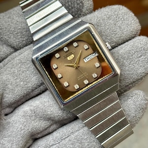 May include: A vintage Seiko automatic wristwatch with a rectangular face. The watch features a brown dial with diamond hour markers and a day-date display. The stainless steel band and case have a brushed finish.
