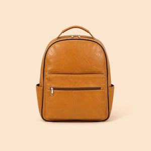 Puede incluir: Una mochila de cuero color canela con un asa superior redondeada, un compartimento principal grande con cierre de doble cremallera y un bolsillo exterior frontal con cremallera horizontal. Incluye dos bolsillos laterales y ribetes oscuros.