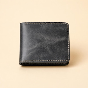 May include: A bi-fold wallet crafted from dark charcoal gray leather with a distressed, weathered texture. The wallet features rounded corners and is constructed with visible, light-colored stitching along the edges, creating a clean, structured rectangular silhouette.
