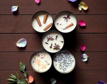 May include: Overhead shot of five small, round candles in silver tins, each with a different topping. Cinnamon sticks, star anise, cloves, and flower petals adorn the creamy wax. The candles are arranged on a dark brown wooden surface.