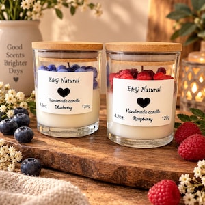 Może przedstawiać: Dwie świece w przezroczystym szkle z bambusowymi wieczkami. Lewa świeca ma etykietę E&G Natural Handmade candle Blueberry 4.6oz 130g i jest zwieńczona niebieskimi jagodami z wosku. Prawa świeca ma etykietę E&G Natural Handmade candle Raspberry 4.2oz 120g i czerwone jagody.