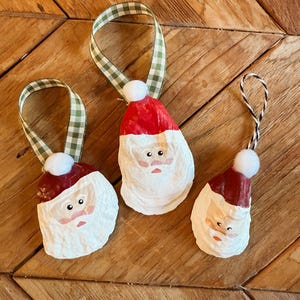 Könnte beinhalten: Drei handgefertigte Weihnachtsschmuckstücke in Form des Weihnachtsmanns. Jedes Ornament hat eine rote Mütze, einen weißen Bart und einen kleinen weißen Bommel. Die Ornamente sind mit grün-weiß karierten Bändern aufgehängt.