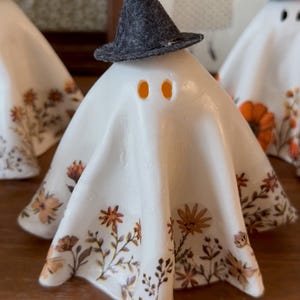 May include: White ceramic ghost figurine wearing a gray felt witch hat. The ghost is decorated with orange and brown floral patterns. The eyes are orange. A Halloween-themed decorative item.