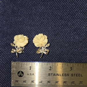 May include: A pair of vintage cream-colored rose earrings. Each earring features a carved rose design with silver-tone accents and a small bow. The earrings are displayed on a blue denim surface next to a ruler.