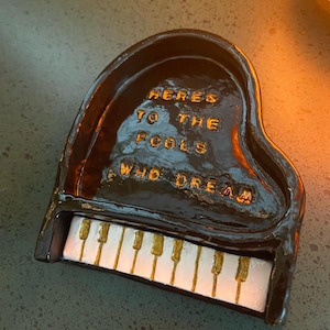 Puede incluir: Un pequeño plato de cerámica en forma de piano, marrón oscuro, con texto dorado que dice "Here's to the fools who dream". Las teclas del piano son blancas y doradas. El plato está sobre una superficie verde oscuro.