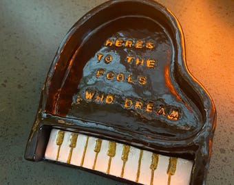 Puede incluir: Un pequeño plato de cerámica en forma de piano, marrón oscuro, con texto dorado que dice "Here's to the fools who dream". Las teclas del piano son blancas y doradas. El plato está sobre una superficie verde oscuro.