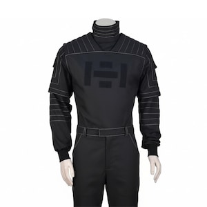 Pu&ograve; includere: Tuta nera con collo alto e maniche lunghe. L'outfit presenta dettagli di cuciture bianche e una cintura. Un grande emblema scuro &egrave; centrato sul petto. Le maniche hanno un design a strati.