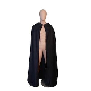 Puede incluir: Una capa negra con cuello redondo y cierre de broche. La capa está drapeada sobre un maniquí, mostrando su diseño fluido. La tela parece ser un material liso y sólido.