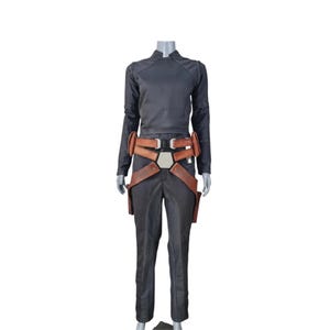 K&ouml;nnte beinhalten: Ein schwarzes Kost&uuml;m, bestehend aus einem lang&auml;rmeligen Oberteil, einer Hose und einem braunen Lederg&uuml;rtel mit Taschen. Das Oberteil hat einen Stehkragen. Der G&uuml;rtel hat eine silberne Schnalle. Das Kost&uuml;m wird auf einer Schaufensterpuppe pr&auml;sentiert.