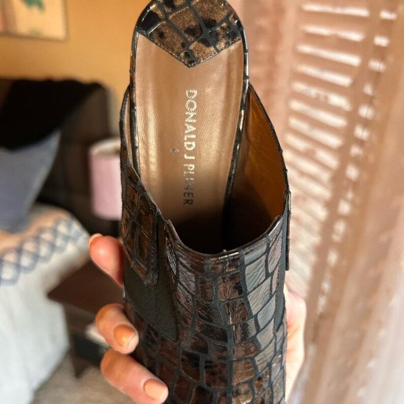 K&ouml;nnte beinhalten: Nahaufnahme einer braunen und schwarzen Maultier-Sandale aus Kunstkrokodilleder. Der Schuh hat eine spitze Zehenpartie und einen kleinen Absatz. Der Markenname "DONALD J PLINER" ist auf der Innensohle zu sehen.