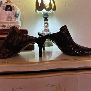 K&ouml;nnte beinhalten: Ein Paar braune Mules mit Absatz und einem strukturierten, krokodilartigen Muster. Die Schuhe haben eine spitze Zehenpartie und ein schwarzes Gummiband oben. Die Abs&auml;tze sind d&uuml;nn und schwarz.