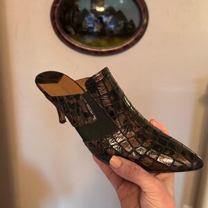 K&ouml;nnte beinhalten: Braune und schwarze Maultier mit Absatz und Krokodilmuster. Der Schuh hat eine spitze Zehenpartie und einen kleinen Absatz. Der Schuh wird von einer Hand gehalten.