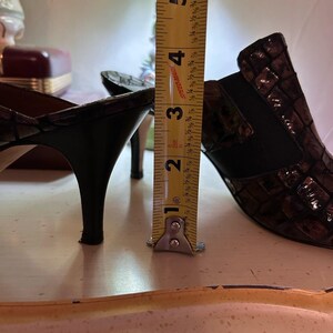 K&ouml;nnte beinhalten: Braune und schwarze Schuhe mit Absatz und strukturiertem Muster. Die Absatzh&ouml;he betr&auml;gt ca. 7,6 cm. Die Schuhe stehen auf einer hellen Oberfl&auml;che.