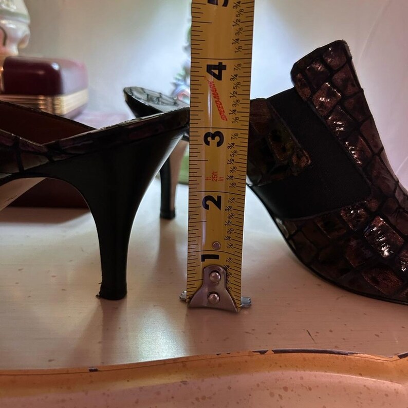 K&ouml;nnte beinhalten: Braune und schwarze Schuhe mit Absatz und strukturiertem Muster. Die Absatzh&ouml;he betr&auml;gt ca. 7,6 cm. Die Schuhe stehen auf einer hellen Oberfl&auml;che.