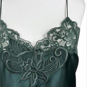 May include: A dark green camisole featuring a V-neckline with intricate floral lace trim. The garment is crafted from a smooth, satin-like fabric and includes thin spaghetti straps. The lace detailing extends across the upper chest area.