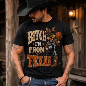 May include: Black t-shirt with the text "BITCH I'M FROM TEXAS" in brown and tan lettering. A cartoon coyote wearing a cowboy hat and Spurs jersey holds a basketball. The coyote is wearing cowboy boots.
