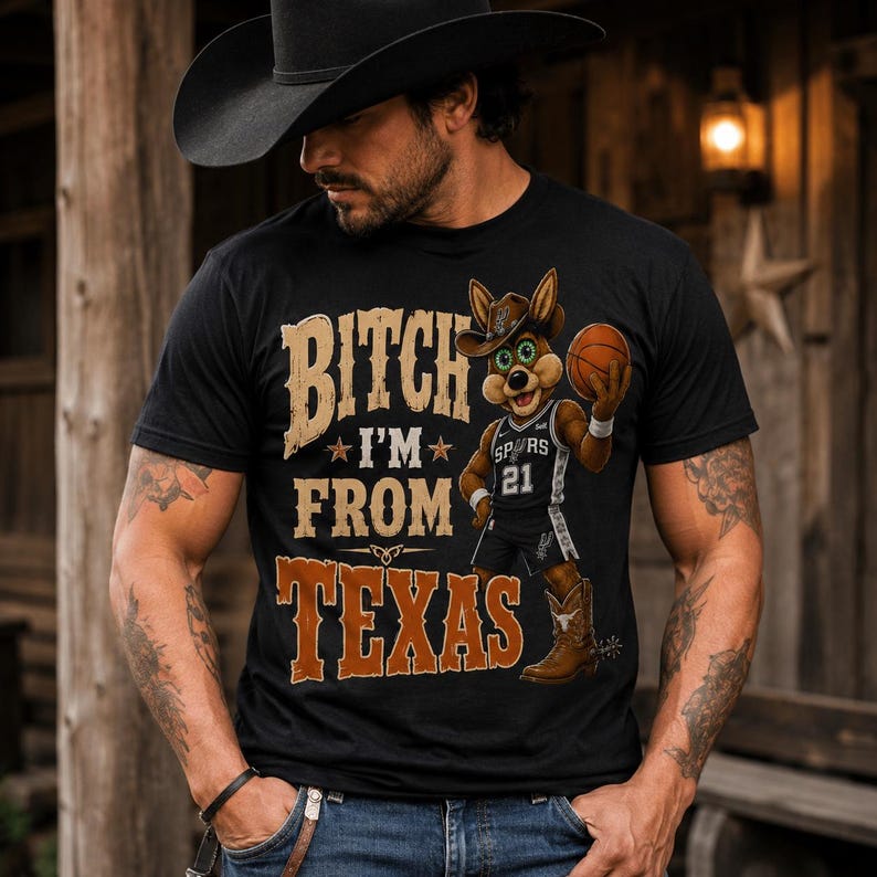May include: Black t-shirt with the text "BITCH I'M FROM TEXAS" in brown and tan lettering. A cartoon coyote wearing a cowboy hat and Spurs jersey holds a basketball. The coyote is wearing cowboy boots.