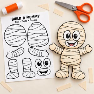 May include: A paper craft activity sheet titled BUILD A MUMMY Cut Paste Create with black and white outlines of mummy body parts, alongside a fully assembled mummy figure with beige bandages, large eyes, and a smiling mouth, plus orange scissors.