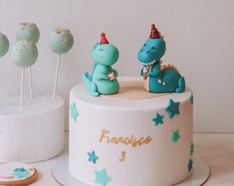 May include: A white birthday cake decorated with teal stars and two dinosaur figurines wearing party hats. The cake has the name "Francisco" and the number "3" in gold lettering. Cake pops and cookies are also present.