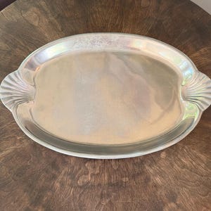 May include: A silver-colored oval serving tray with shell-shaped handles. The tray has a reflective surface and is set on a dark wooden table. The tray is empty and ready for use.
