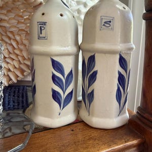 May include: A pair of ceramic salt and pepper shakers. The shakers are white with blue leaf designs and the letters "P" and "S" in blue. The salt and pepper shakers are on a wooden surface.