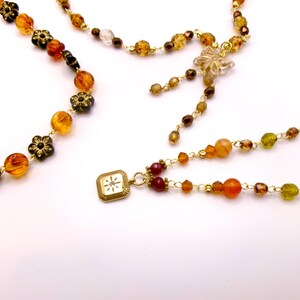 May include: Close-up of several necklaces with amber, brown, and clear beads. Some necklaces feature flower-shaped accents and a gold-colored square pendant with a starburst design. The necklaces are on a white background.