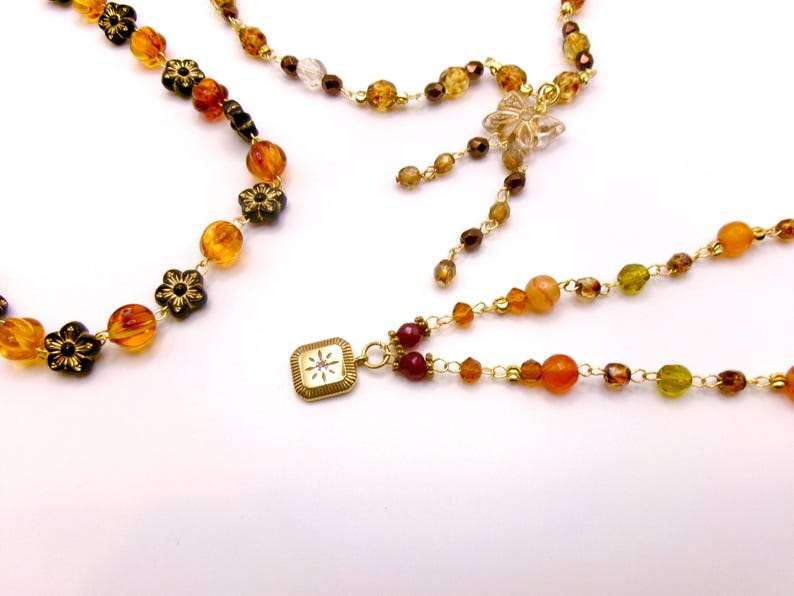 May include: Close-up of several necklaces with amber, brown, and clear beads. Some necklaces feature flower-shaped accents and a gold-colored square pendant with a starburst design. The necklaces are on a white background.