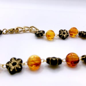 May include: A gold-toned necklace featuring alternating amber-colored beads and black flower-shaped beads. The necklace has a gold chain clasp. The beads are strung on a delicate gold chain.