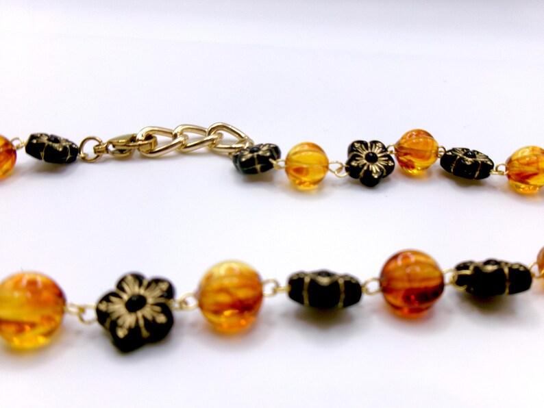 May include: A gold-toned necklace featuring alternating amber-colored beads and black flower-shaped beads. The necklace has a gold chain clasp. The beads are strung on a delicate gold chain.