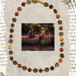 May include: A necklace featuring amber-colored beads and gold-toned flower accents. The necklace surrounds a small image of two people sitting on a wooden structure near water. The necklace has a gold clasp.