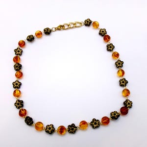 May include: A necklace featuring amber-colored beads and black flower-shaped accents. The necklace has a gold-colored chain and clasp. The beads are round and vary in color from light to dark amber. The flower accents are black with intricate details.