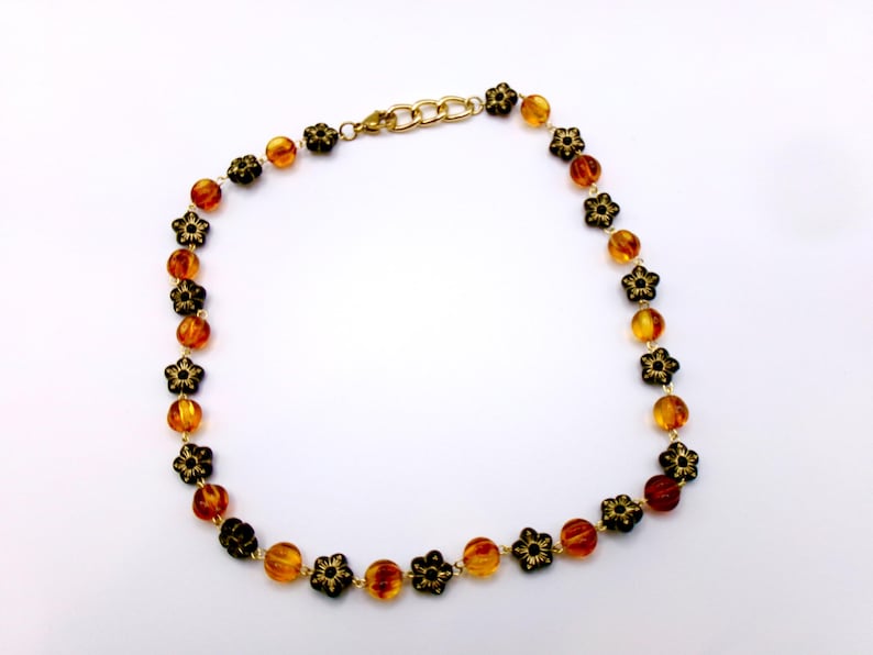 May include: A necklace featuring amber-colored beads and black flower-shaped accents. The necklace has a gold-colored chain and clasp. The beads are round and vary in color from light to dark amber. The flower accents are black with intricate details.