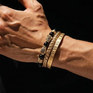Könnte beinhalten: Drei goldene Armbänder an einem Handgelenk. Eines hat schwarze Perlen und goldene Akzente, ein anderes ein geflochtenes Design und das dritte ist ein goldener Armreif mit römischen Ziffern. Die Armbänder sind übereinander gestapelt.