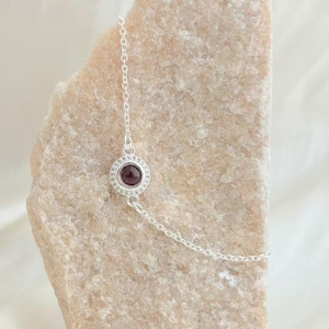 Könnte beinhalten: Ein silberfarbenes Handkettchen-Armband mit einem kreisförmigen Anhänger mit einem dunkelroten Edelstein in der Mitte, umgeben von kleinen, klaren Steinen. Das Armband wird auf einem hellbeige, strukturierten Stein präsentiert.