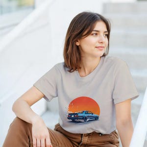 May include: A light gray t-shirt featuring a blue car graphic with a red and orange sunset design. The car is depicted with smoke trails, suggesting speed. The shirt is worn by a person.