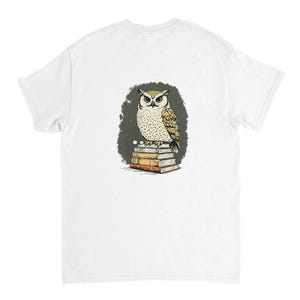 Könnte beinhalten: Ein weißes T-Shirt mit einer grafischen Illustration einer beigen und braunen Eule, die auf einem Stapel von vier Büchern sitzt. Die Eule hat detaillierte Federn und sitzt auf den Büchern, die in Beige-, Braun- und Orangetönen gehalten sind.