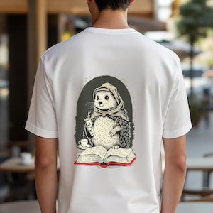 Könnte beinhalten: Ein weißes T-Shirt mit einer Grafik auf der Rückseite, die einen Igel mit Kapuzenumhang zeigt, der eine dampfende Tasse und eine Schriftrolle vor einem aufgeschlagenen Buch hält. Die Illustration ist in Braun-, Grau- und Rottönen gehalten.