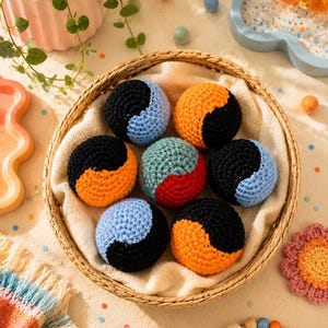 May include: A woven basket holds seven crocheted balls in a variety of colors, including black, orange, blue, and green. The balls are arranged on a white cloth. The basket is surrounded by other decorative items, including a plant and a colorful blanket.