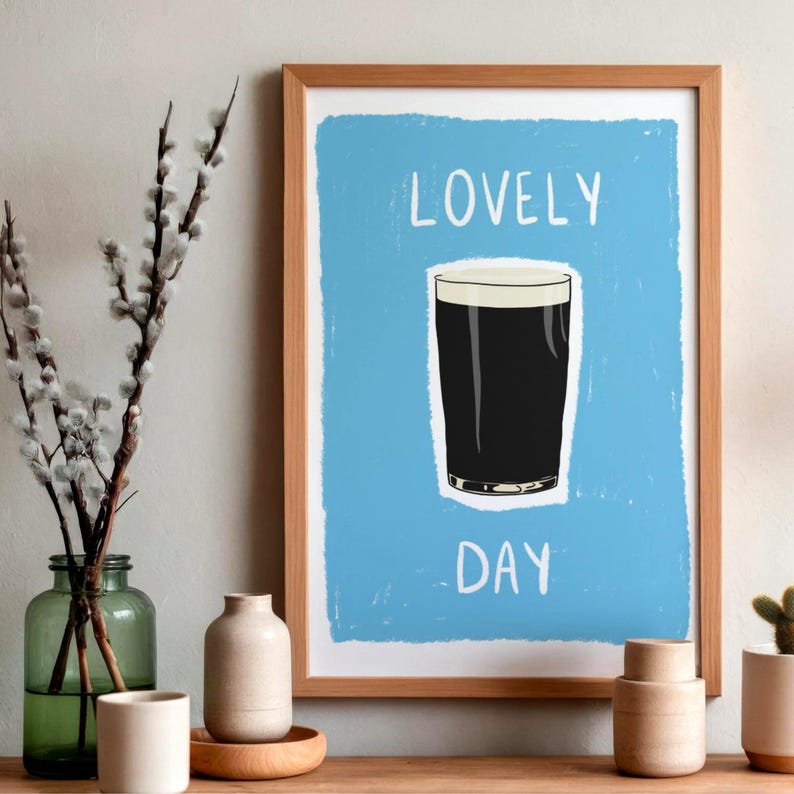 May include: A framed wall art print featuring a light blue background with a dark stout beer in a glass centered between the white text LOVELY and DAY. The print is enclosed in a light wood frame, showcasing a clean and simple graphic design.